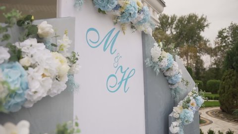 Wedding arch in the park among nature, trees, grass and flower beds. Wedding day. Preparation for the ceremony. Floral arrangements adorn the arch. The letters "M" and "H" are written on the arch