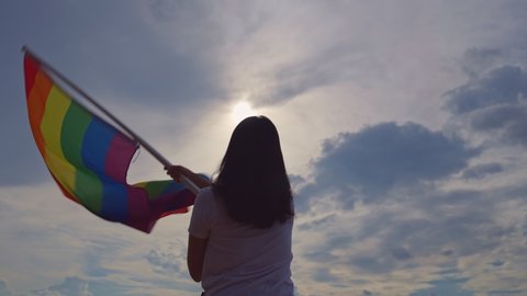 Slow motion 4K. Woman holding lgbtq flag in hand. Female waving rainbow LGBT flag on sunset blue sky with pride. Lesbian, Gay, Bisexual, transgender social movements. Freedom of love concept.