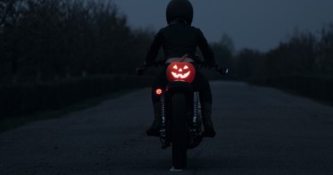 Halloween pumpkin lantern rides on the back seat of a biker motorcycle, biker rides a dark road into the distance with a glowing halloween lantern