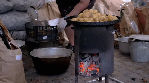 Street Chef Prepares Potatoes on a Wood Stove with Glowing Coals. Men shifts washed potatoes in a box made of cast iron cauldron. Cooking of grilled vegetables with steam and smoke. Slow motion.
