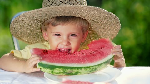 Handsome toddler boy eating fresh red watermelon. Yummy footage of smiling child sits by the table in garden. Healthy lifestyle, melon vitamins, summer harvest.