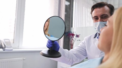 Back view male professional dentist doctor man shows mirror reflection to young satisfied woman patient dental clinic light office medical center with modern tools equipment. Healthy lifestyle concept