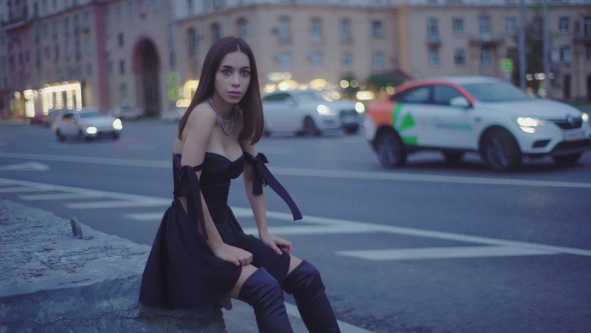 Romantic stylish woman in silk black dress sitting in evening city. Affectionate brunette woman posing near night highway with moving cars. Attractive Appearance. 4K
