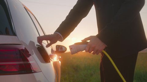 Handheld shot of a Caucasian businessman in a black suit charging an electric car at a charging station near a highway at sunset, using his smartphone while waiting