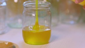 Unknown woman pours sweet golden honey into transparent empty clean jars standing on a wooden white large table on a bright sunny day, 4K UHD slow-motion video