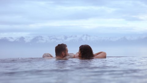 REYKJAVIK, ICELAND - JUNE 2021: Beautiful still shot of a couple intimately staring at each other, bathing in an infinity pool with the landscape of the majestic mountains with snow