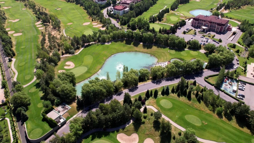 A golf course with a view of the high mountains. Blue water in the pond, green grass and places to serve the ball. Tall coniferous trees, blue sky and houses. The view from drone. Kazakhstan, Almaty