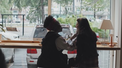Rear view slowmo of multi-ethnic lesbian couple sitting at table by window in cafe and enjoying food. Caring African-American woman wiping mouth of her Caucasian girlfriend