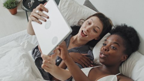 High angle handheld slowmo shot of happy multi-ethnic lesbian couple lying in bed and laughing while chatting and looking at tablet