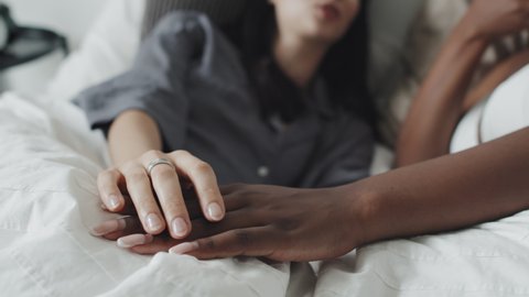 Handheld close up of Caucasian young woman lying in bed and stroking hand of her African-American female partner while chatting and relaxing in morning