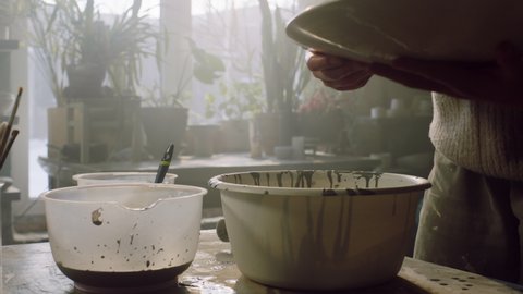 Woman ceramist is pouring glaze on big plate, glazing it manually, rotating the plate to spread liquid evenly, ceramics art, Foreground, Slow motion.
