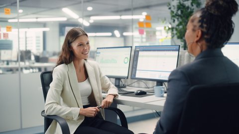 Two Female Colleagues Fondly Talk to Each Other, Laugh and Smile while Working on Computers in Diverse Modern Business Office. Experienced Manager and Young Employee Discuss a Fun Analytical Project.