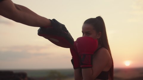 Woman in boxing gloves trains with coach in paws nature at sunset, front view. Girl trains to box with trainer the evening nature