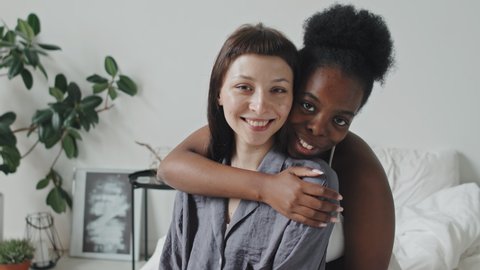 Handheld slowmo portrait of happy African-American woman finishing brushing her Caucasian girlfriends hair and hugging her with affection. They are looking at camera and smiling