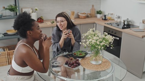Slowmo shot of beautiful lesbian couple sitting at kitchen table and chatting while enjoying coffee in morning