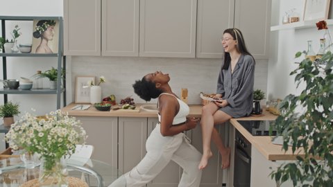 Slowmo medium shot of happy African-American woman throwing plum and catching it with her mouth while her Caucasian girlfriend sitting on kitchen counter and laughing