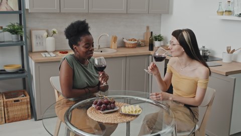 Handheld slowmo of happy African-American woman and her Caucasian girlfriend sitting at round glass table in cozy kitchen and drinking wine
