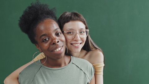 Portrait close up of multi-ethnic female couple hugging and smiling for camera while posing against dark green background