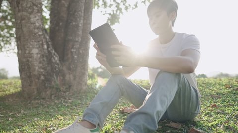 Young asian man sitting under big tree in park with hands holding digital laptop, freelancer in casual clothing remote working outdoor, sit on comfortable meadow with lens flare behind, nature people