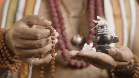 Close up hands of Indian priest meditating or chanting using rudrakshi japa mala with shiva linga in hand