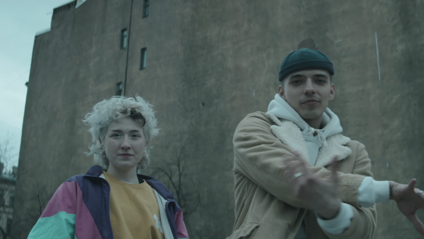 Waist up of young stylish couple looking at camera and performing vogue dance outdoors against old concrete building