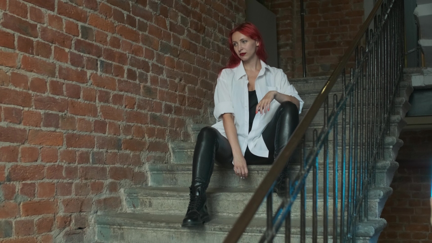A beautiful red-haired girl wearing leather trousers, belts, black and white top and ring in her nose looking straight sitting on stairs in red brick building or block of flats Royalty-Free Stock Footage #1078203416