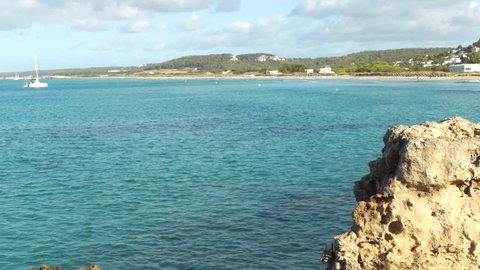 Wide Distant view of Son Bou beach the longest in Menorca, clear and calm water with boats, Balearic Islands, Spain. Rock in foreground.