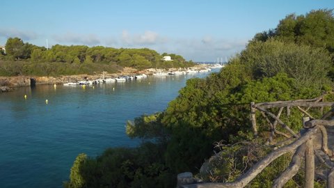 Wide view from a high point behind the wooden barriers of the Cala Santandria in Menorca just after dawn, blue seawater, surrounding rocks, boats, green trees.