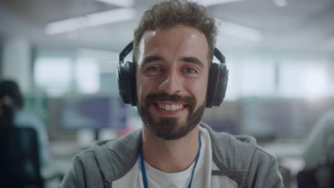 Office: Portrait of Happy IT Programmer Wearing Headphones Working on Desktop Computer, Looking at Camera and Smiling. Male Software Engineer Developing App, Video Game. Listening to Podcast, Music