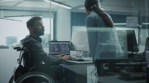 Disability-Friendly Office: Brilliant IT Programmer with Disability in a Wheelchair Talks with Colleague while Working on Computer. Software Engineers Develop Innovative App, Program. Medium Shot