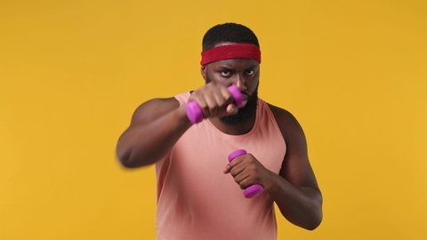 Serious strong slim trainer instructor young african american man sportsman 20s in headband pink tank top warm up train exercise with dumbbells in home gym isolated on plain yellow background studio