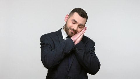 Tired bearded businessman in black official style suit feeling drowsiness, it is late and falling asleep, keeping palms together near ear. Indoor studio shot isolated on gray background.