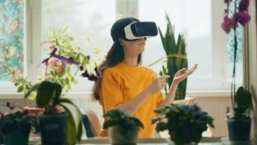 Young pretty Caucasian woman in VR glasses painting with virtual brush. Indoor, interior with natural plants. Window in the background. The concept of futururistic technologies.