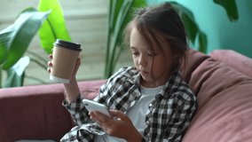 Close-up of amazed little child girl scrolling phone screen and saying wow sitting in armchair holding coffee cup at home. Pretty young girl with long blond hair using smartphone and looking surprised