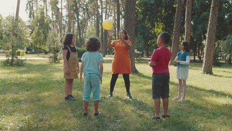 Group of multiracial diverse school kids playing ball with lovely young female teacher in public park during outdoor classes at morning. Leisure activity of charming children having fun after lesson