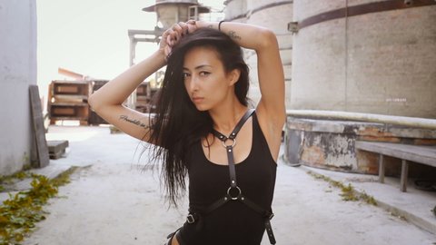 Close-up of sexy Asian girl in black swimsuit and leather harness smiling and posing in abandoned industrial zone. Go-go show at techno festival. Slide camera. Cyberpunk. Fetish military. BDSM