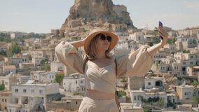 Young pretty woman tourist in a hat and with a backpack makes a selfie on the background of the old city with Ortahisar fortress, Travel in Turkey, Cappadocia