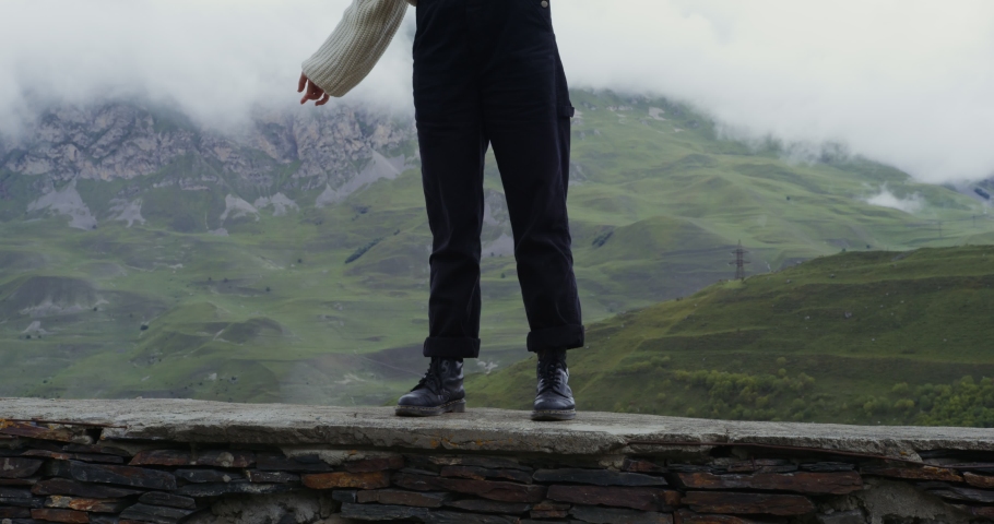 Russia, Caucasus. A beautiful young woman of European appearance, dressed in a warm sweater, stands on the parapet against the background of green hills and makes smooth movements with her hands