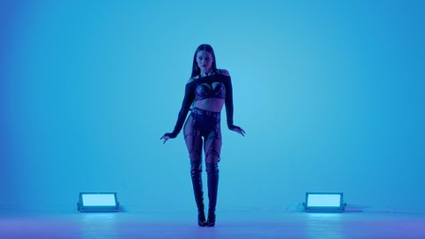 Sexy brunette in black leather lingerie is dancing erotically in a dark studio illuminated by blue lights. Sensual stripper dance, private dance, go go. Slow motion.