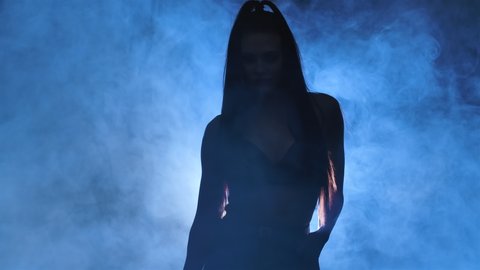 Silhouette of sexy brunette in black leather lingerie is dancing erotically in a dark smoky studio illuminated by blue lights. Sensual stripper dance, private dance, go go. Close up. Slow motion.
