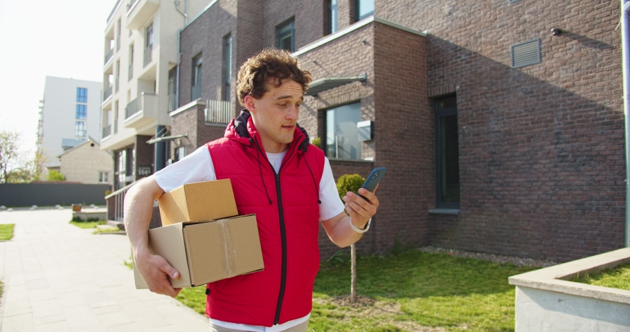 Caucasian male delivery worker going through the street and carrying cupboard boxes with goods while looking at smartphone. Male courier searching route to the client