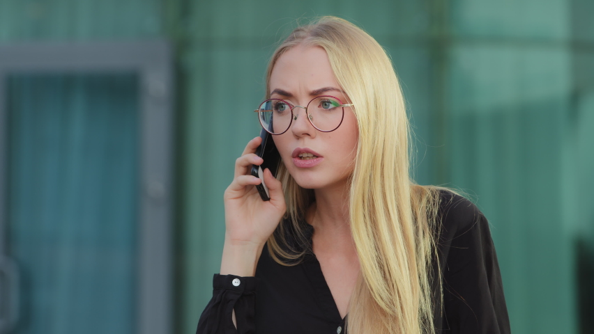 Worried nervous Caucasian European blonde woman holding mobile phone angrily talking to bank service outdoors. Customer dissatisfied with poor serve, annoyed client, aggressive businesswoman concept