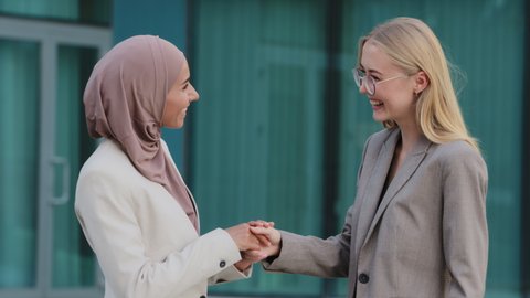 Happy diverse millennial female colleagues or partners get acquainted greeting each other by handshaking glad to meet. Smiling businesswomen Indian girl in hijab and young European woman shaking hands