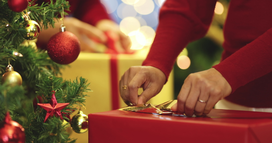 Hands of two women in red dress holding and using ribbon to tie bow for a wrapped gift box for Christmas present with Xmas tree in foreground and colorful lights bokeh in blur background. | Shutterstock HD Video #1078273898