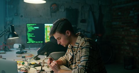 Man is Soldering in Electronics Workshop. Hacking Crime, Cyber War or IT Startup Concept. Young Coders Work on Technology Project in Dark Garage, Basement or Loft Office. 4K Wide Pan Right shot