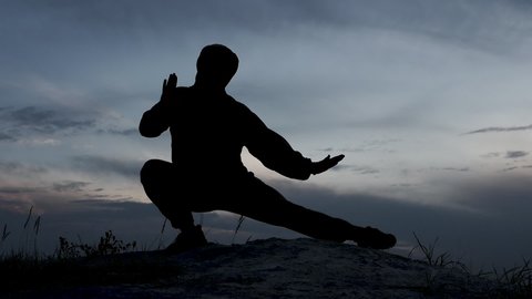 Silhouette of a man at sunset on the top of a mountain, a man practicing the art of Shaolin martial arts, a sports fighter training in nature. The young man is studying martial arts.