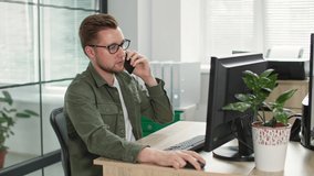 young businessman with glasses talking on phone while working at computer in office background of window
