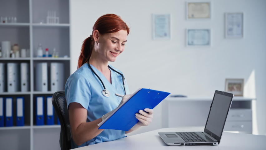 portrait of young female doctor in medical uniform during an appointment with patient online using video call on laptop while sitting in clinic office, smiling and lookin