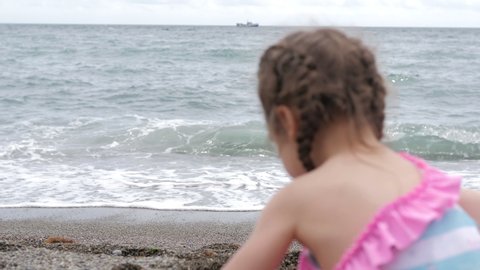 A little 3-year-old girl is sitting by the sea on a bright sunny day and playing with pebbles on a pebble beach. Relaxing on the beach. Sandy beach. Sea air. A child plays by the sea