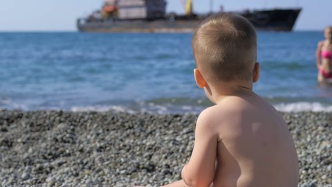 Close-up, A small one-year-old boy is sitting by the sea on a bright sunny day and playing with pebbles on a pebble beach. Relaxing on the beach. Sandy beach. Sea air. A child plays by the sea.